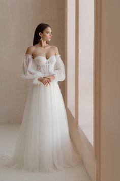 a woman standing in front of a window wearing a white wedding dress with sleeves on it