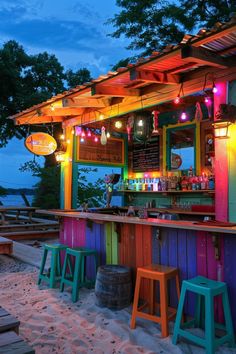 an outdoor bar with colorful lights and stools on the sand at dusk, next to trees