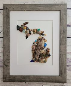 a bird made out of rocks and pebbles in a white frame on a wooden wall