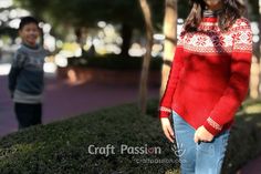 two people standing next to each other in front of some bushes and trees with one person wearing a red sweater