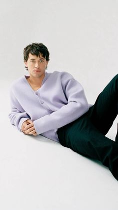 a man sitting on the ground wearing a purple sweater and black pants with his legs crossed