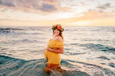 a pregnant woman standing in the ocean at sunset wearing a wreath of flowers on her head