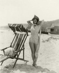 a woman in a bathing suit and hat standing on the beach next to a chair