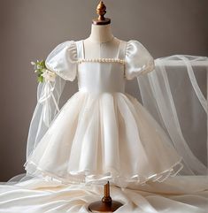 This charming flower girl and communion dress combines timeless elegance with whimsical charm. Expertly tailored to order, this dress features delicate puff sleeves and a layered tulle skirt that catch the light beautifully. The square neckline is beautifully embellished with a pearl trim, mirroring the sophistication of bridal designs. Perfect for weddings, communions, or any formal event where your little one needs to shine. This custom-fit gown is designed to provide a perfect, comfortable fi Elegant Princess Dress With Ruffles For First Communion, Elegant Cream Princess Dress With Tulle Skirt, Elegant Cream Tulle Princess Dress, Organza Puff Sleeve Wedding Dress, Elegant Ruffled Tutu Dress For First Communion, First Communion Princess Dress With Ruffles, Tulle First Communion Dress With Ruffles For Wedding, Elegant Organza Puff Sleeve Dress For Wedding, Elegant Organza Puff Sleeve Wedding Dress