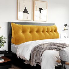 a bed with yellow tufted headboard and white sheets in a small room next to two framed pictures on the wall