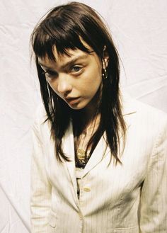 a woman with long hair wearing a white suit and gold earrings, standing in front of a white backdrop