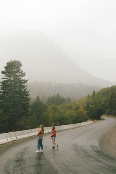 two people walking down the road on a foggy day with mountains in the background