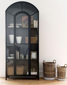 an arched glass cabinet with wicker baskets on the floor next to it and a white wall in the background