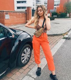 a woman standing next to a car talking on a cell phone while wearing an orange pantsuit