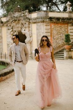 a man and woman walking in front of an old building with stone steps, one wearing a pink dress