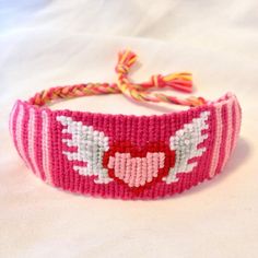 a pink bracelet with white wings and a red heart on it's front end