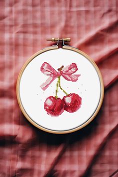 a cross - stitch cherry with two bows is hanging on a piece of red fabric