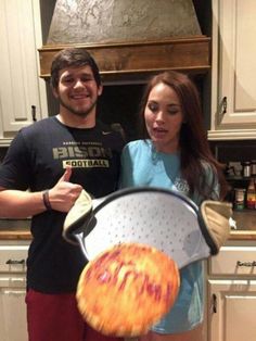 a man and woman standing in a kitchen holding a pizza pan shaped like a face