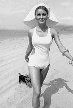 a woman in a bathing suit and hat walking on the beach with her arm outstretched