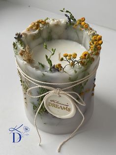 a white candle with yellow flowers on it sitting in a glass container next to a string