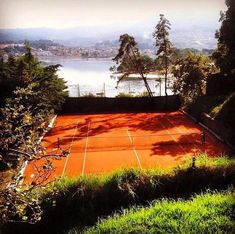 an orange tennis court in the middle of some trees and grass with water in the background