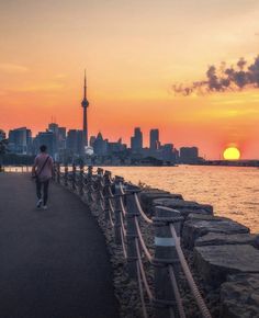 a person walking down a path near the water with buildings in the background at sunset