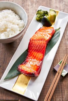 a white plate topped with fish next to rice and chopsticks