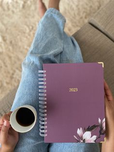 a person sitting on the floor holding a cup of coffee and a purple planner book