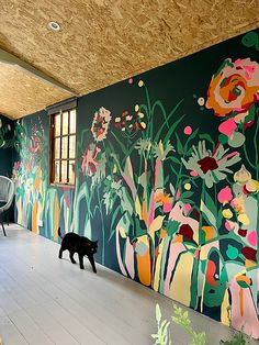 a black cat is walking in front of a colorful wall with flowers and plants on it