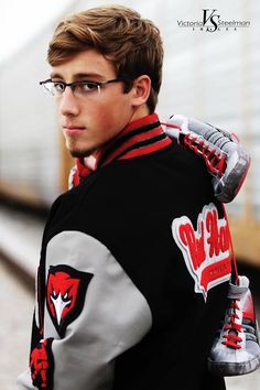 a young man wearing glasses and a jacket with patches on the sleeves is standing in front of a train