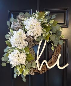 a wreath with the letter h on it is hanging on a black door and has white flowers