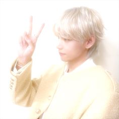 a person with blonde hair making the v sign in front of a white wall and holding their hand up