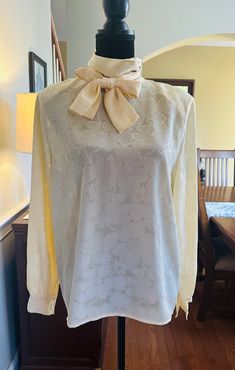 A vintage long sleeve blouse by Kira Evans.  Lightweight ivory polyester fabric with a embossed floral design.  High neck, with 2 button and hook closure at the back of the neck.  Attached tie that can we worn various ways.  The bust measures 20 inches from armpit to armpit laying flat.  24.5 inches from shoulder to hem.  No rips or stains.  Please examine the pics, feel free to ask questions. Womens Blouses, Vintage Long Sleeve, We Wear, Beautiful Dolls, Womens Clothing Tops, Etsy Vintage, Sleeve Blouse, Polyester Fabric, High Neck