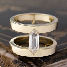 two gold wedding rings with an emerald cut diamond on each one, sitting on a piece of wood