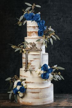 a three tiered cake with blue flowers and greenery on the top is displayed in front of a black background