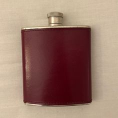 a red leather flask with a silver top on a white cloth covered wall behind it