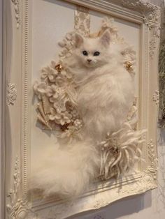 a white cat is hanging on the wall in front of a frame with flowers and feathers