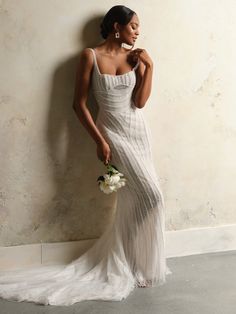 a woman in a white wedding dress leaning against a wall holding a bouquet and looking off to the side