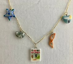 Vintage-themed charm necklace on a gold bar chain; all the charms are sculpted from a fine white porcelain clay and painted by hand to have charming details. Back and front sides are painted. Since they are made from porcelain and glossed with a clear glaze, each charm is smooth and soft to the touch.  Each charm is attached to a small, gold jump ring and can be attached to a necklace chain. Small charms featured: New Yorker cover with cat looking out at city, Curled up cat sleeping, Green Star Fish, Blue Star Fish, Love Letter w/ gold highlights, Red Lipstick with gold band, Red lips, Slipper with gold heart, Gold heart (gold raindrops on one side, gold heart on the other), Lucky Strikes pack, Moon and Black Cat on a heart, Sun Hat with gold band. Bought individually, your charm(s) will c Clay Star Necklace, Diy Initial Necklace, Curled Up Cat, Clay Charm Necklace, Clay Chain, Highlights Red, New Yorker Cover, Lucky Charm Necklace, Green Star