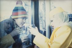 two women are looking at their cell phones in front of a store window, one is wearing a knitted hat and the other wears a yellow coat