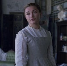 a woman standing in front of a bed wearing a white shirt and dress with ruffled sleeves