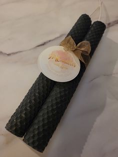 two black candles with a brown bow on them sitting on a marble counter top next to a tag