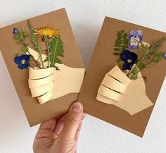 two handmade greeting cards with flowers in vases on brown paper, being held by a person's hand