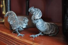 two metal rooster figurines sitting on top of a wooden shelf