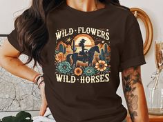 This graphic T-shirt features a unique Wild Flowers Wild Horses design, perfect for anyone who loves vintage western themes and boho styles. Showcasing a retro desert scenery with floral elements and a cowboy rider, it adds a touch of rodeo charm to your wardrobe. Ideal for casual outings or themed events, this shirt is a great addition to any collection.  WELCOME TO RAIN APPAREL US  We are a small business located in Missouri. We started making t-shirts in 2019 and have grown so much since then! We take pride in the quality of our products and are always striving to grow. We hope you can find whatever you are looking for in our shop. Please reach out to our team if you need any help at all! Quality Assurance: We pride ourselves on using high-quality fabrics and safe inks. Our suppliers as Horses Design, Retro Desert, Desert Scenery, Rodeo Style, Boho Desert, Flowers Wild, Maroon Shirts, Cowboy Shirt, Rodeo Fashion