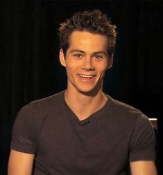 a man smiling while sitting down in front of a black curtain with his hands on his hips