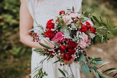 a woman holding a bouquet of red and pink flowers with greenery in her hands