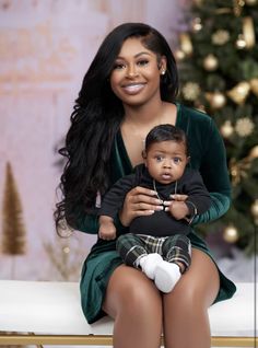 a woman sitting next to a baby in front of a christmas tree holding a doll