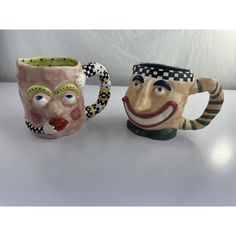 two ceramic mugs with faces on them sitting next to each other, one has a clown's head