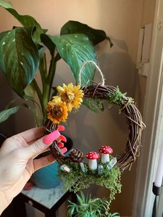 a hand holding a wreath with flowers and mushrooms on it next to a potted plant