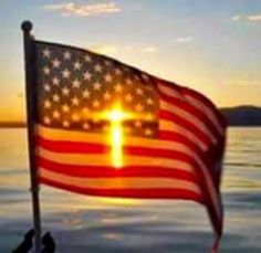 an american flag with a cross on it flying in the air over water at sunset