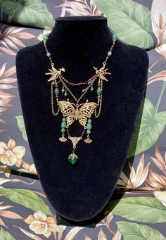 This exquisite necklace boasts a stunning Art Nouveau chandelier style that is sure to turn heads. The centerpiece of the necklace is a beautifully crafted antique bronze butterfly that adds a touch of vintage charm to the piece. The necklace features intricate hanging chains that lend an air of elegance to the design. The gemstones used in this necklace include green adventurine, garnet, and African turquoise, which complement each other perfectly to create a harmonious color scheme. The necklace measures 17 inches in length with a 1 inch extender, making it a perfect fit for a variety of neck sizes. Bohemian Butterfly Jewelry For Festivals, Bohemian Necklace With Butterfly Charm, Bohemian Jewelry With Butterfly Charm Dangle, Bohemian Dangle Jewelry With Butterfly Charm, Handmade Gold Bohemian Butterfly Necklace, Bohemian Butterfly Jewelry, Bohemian Butterfly Charm Jewelry For Jewelry Making, Bohemian Jewelry With Butterfly Charm For Jewelry Making, Gold Bohemian Butterfly Necklace