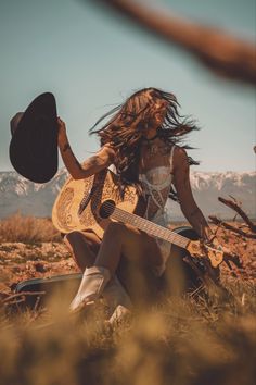 a woman sitting on the ground with her guitar in hand and mountains in the background