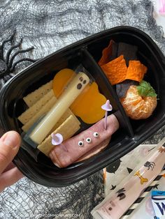 a person is holding a black container with food in it that includes meat, cheese and fruit