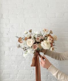 a woman holding a bouquet of flowers in front of a white brick wall with a brown ribbon tied around it